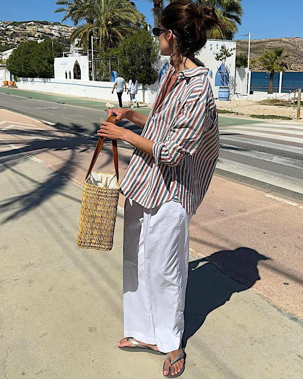 Femme en chemise rayée et pantalon blanc large marchant près de la mer avec un panier en osier