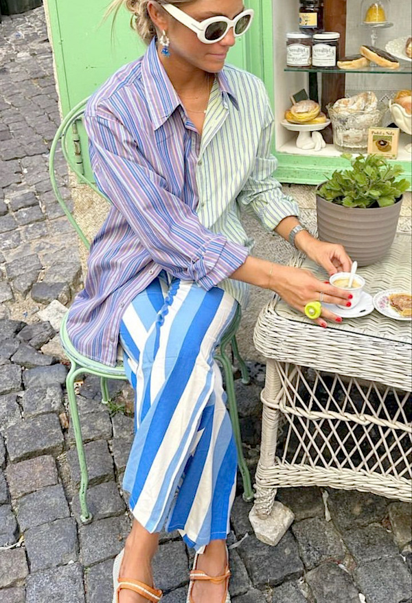 Femme assise à la terrasse d’un café avec une chemise rayée multicolore, un pantalon rayé bleu et blanc et des lunettes blanches